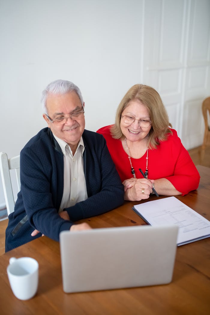 why-choose-us Senior couple sitting at a table, smiling while using a laptop indoors. Perfect for tech and lifestyle themes.