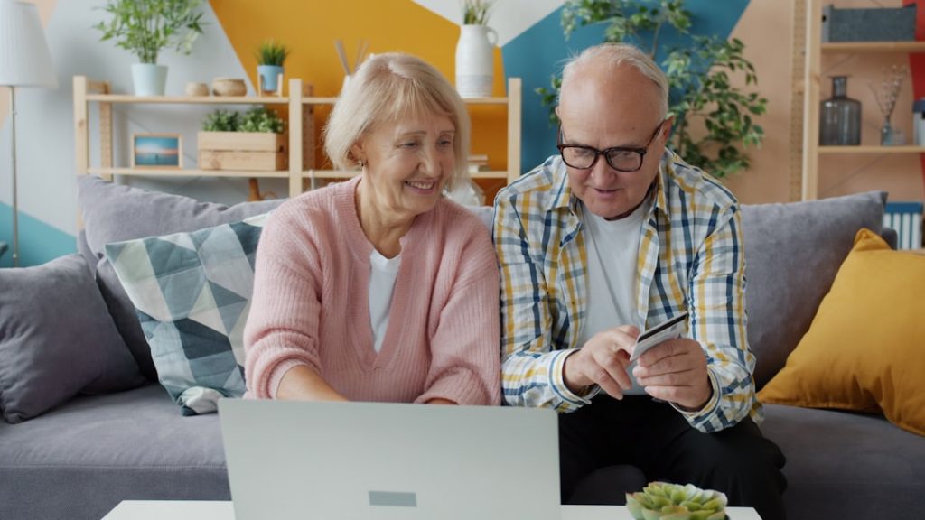 Cheerful elderly people man and woman are making cashless online payment with bank card using laptop at home. Money, retirement and shopping concept.